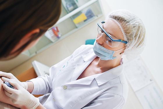 A medical professional wearing a white coat, 眼镜, and medical mask administers a needle to a patient's arm. 